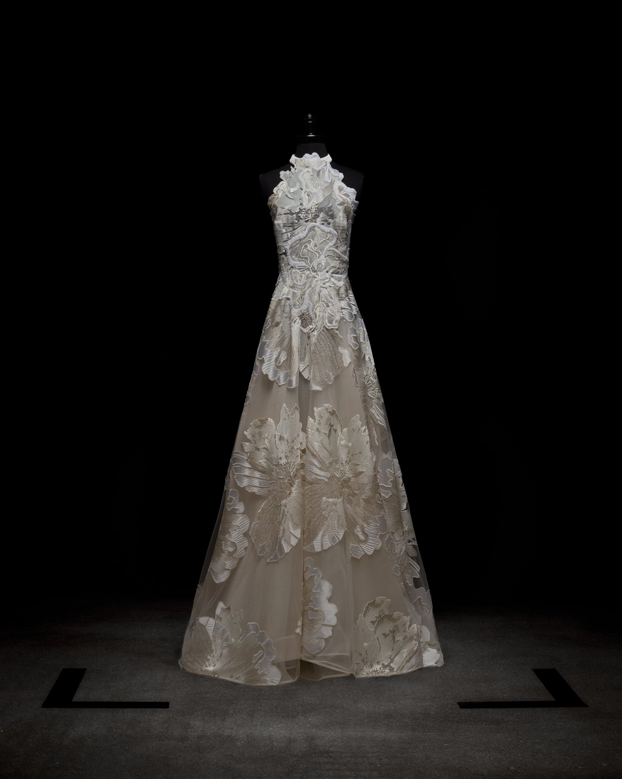 The Becca Dress by SVT DESIGNS, displayed on a mannequin against a dark background, features intricate white floral lace patterns over sheer fabric, creating an elegant and delicate appearance.