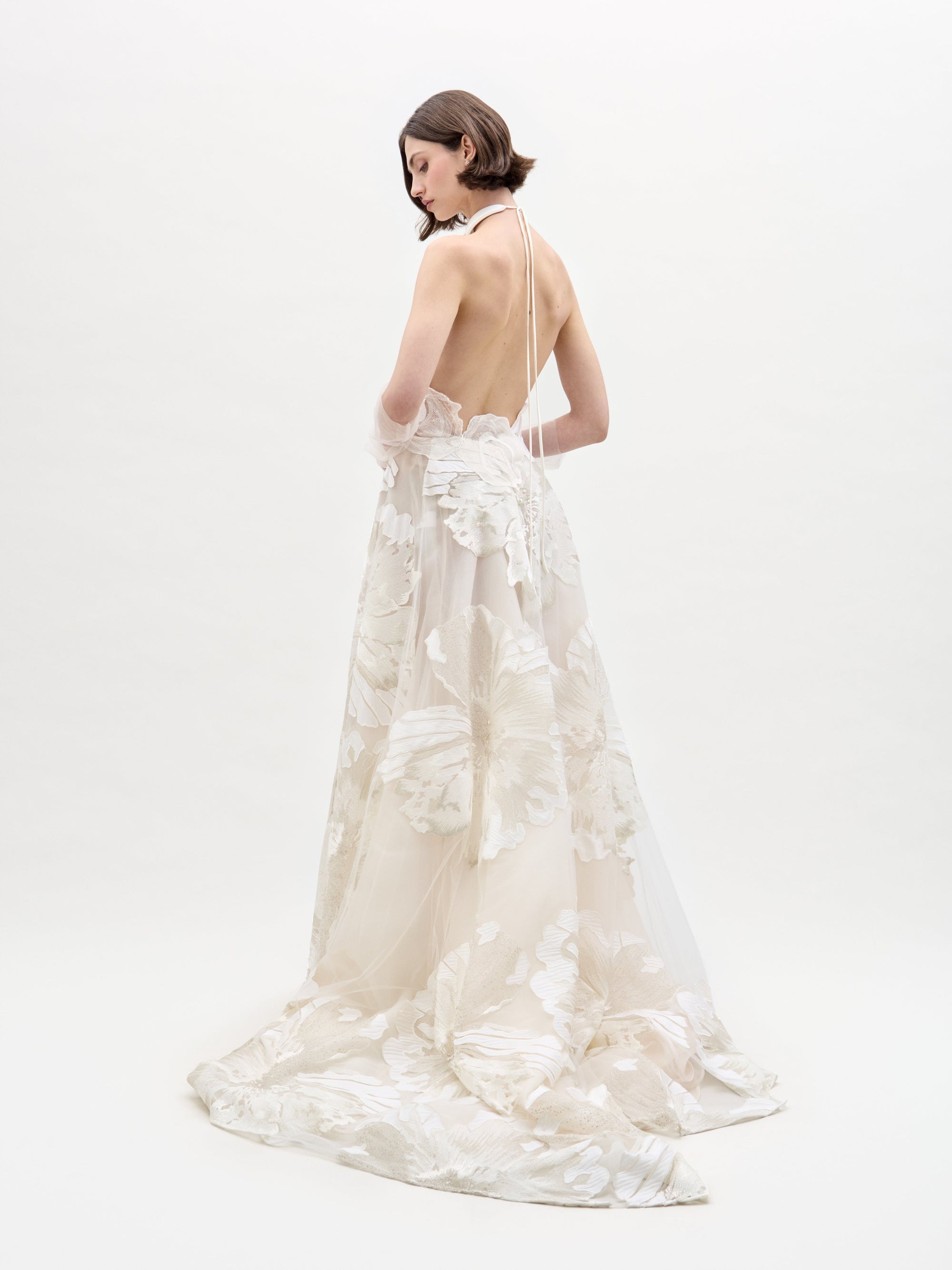 In a studio with a plain white background, a woman with short, straight hair models the Becca Dress by SVT DESIGNS. The elegant white gown features floral patterns, a halter neck, and an open back as she stands facing to the side.