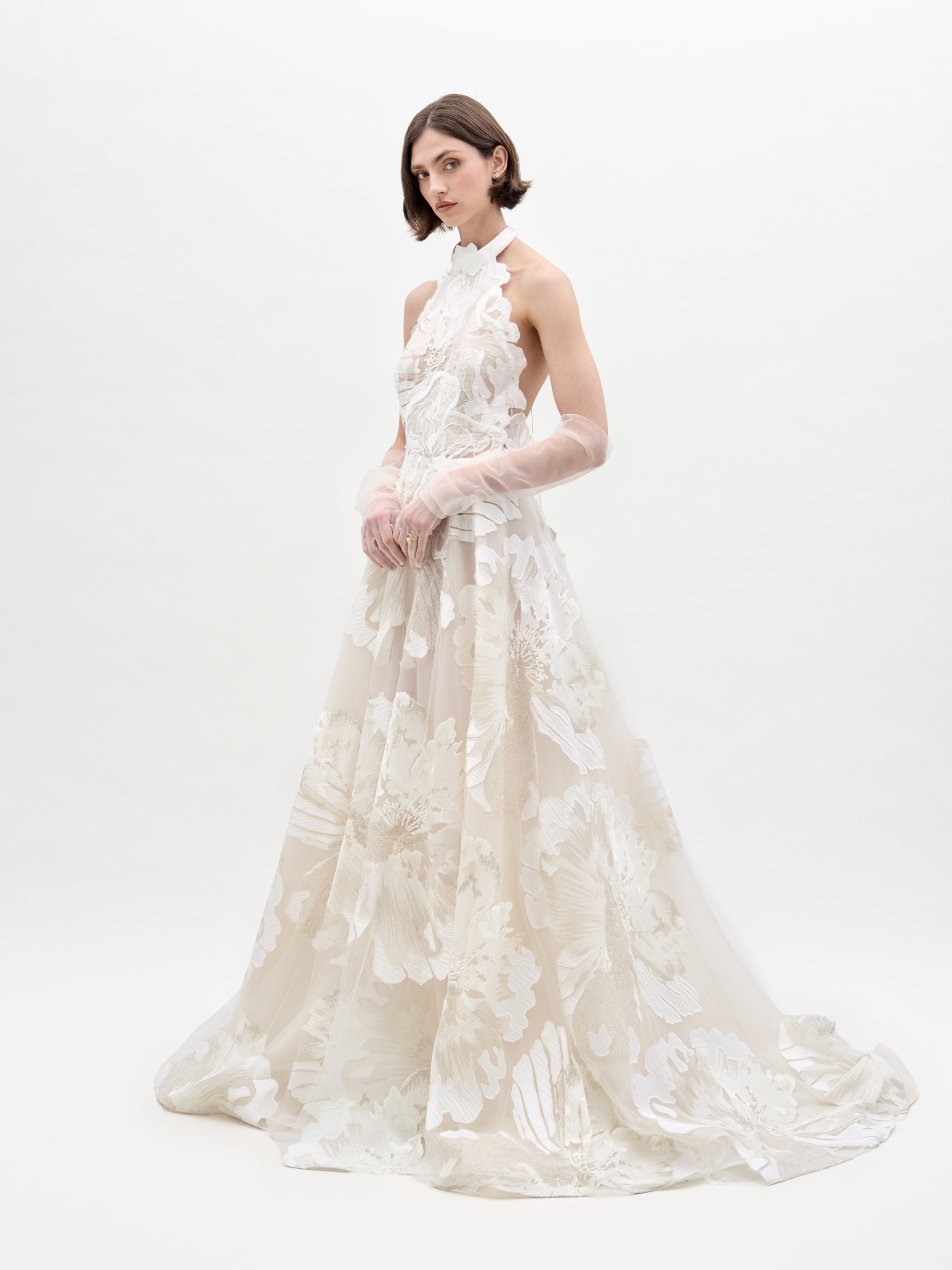 A person wearing the SVT DESIGNS Becca Dress, an elegant floor-length gown with a floral pattern, stands against a light background. The dress has a high neckline and is paired with sheer gloves. They have short hair and maintain a neutral expression.