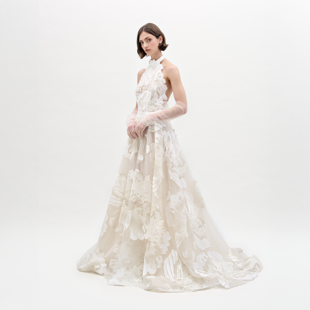 A woman with short brown hair poses against a white background in the SVT DESIGNS Becca Dress, a floor-length, sleeveless white gown featuring large floral patterns in Italian fabric. She pairs it with sheer, elbow-length gloves.