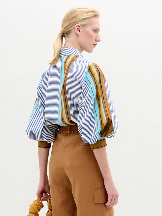 A person with blonde hair in a low ponytail wears an oversized multicolored striped Belkis Blouse by SVT DESIGNS featuring voluminous sleeves and tan trousers. They hold a brown scrunchie in their left hand, turned slightly to the right against a plain background.