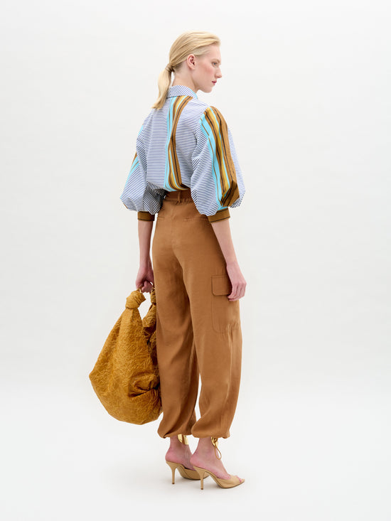 A blonde woman stands against a plain background, looking to the side. She's dressed in a striped blouse, SVT DESIGNS' Darnell Pant, and high heels. In one hand, she carries a large, textured tote bag made from fine Italian fabric.