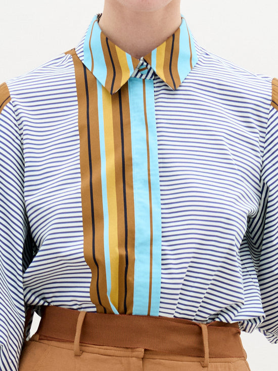 A person in an oversized SVT DESIGNS Belkis Blouse features horizontal blue and white stripes with vertical brown, yellow, and blue stripes. The Italian fabric cotton blouse is tucked into high-waisted tan pants. The head is not visible in the image.