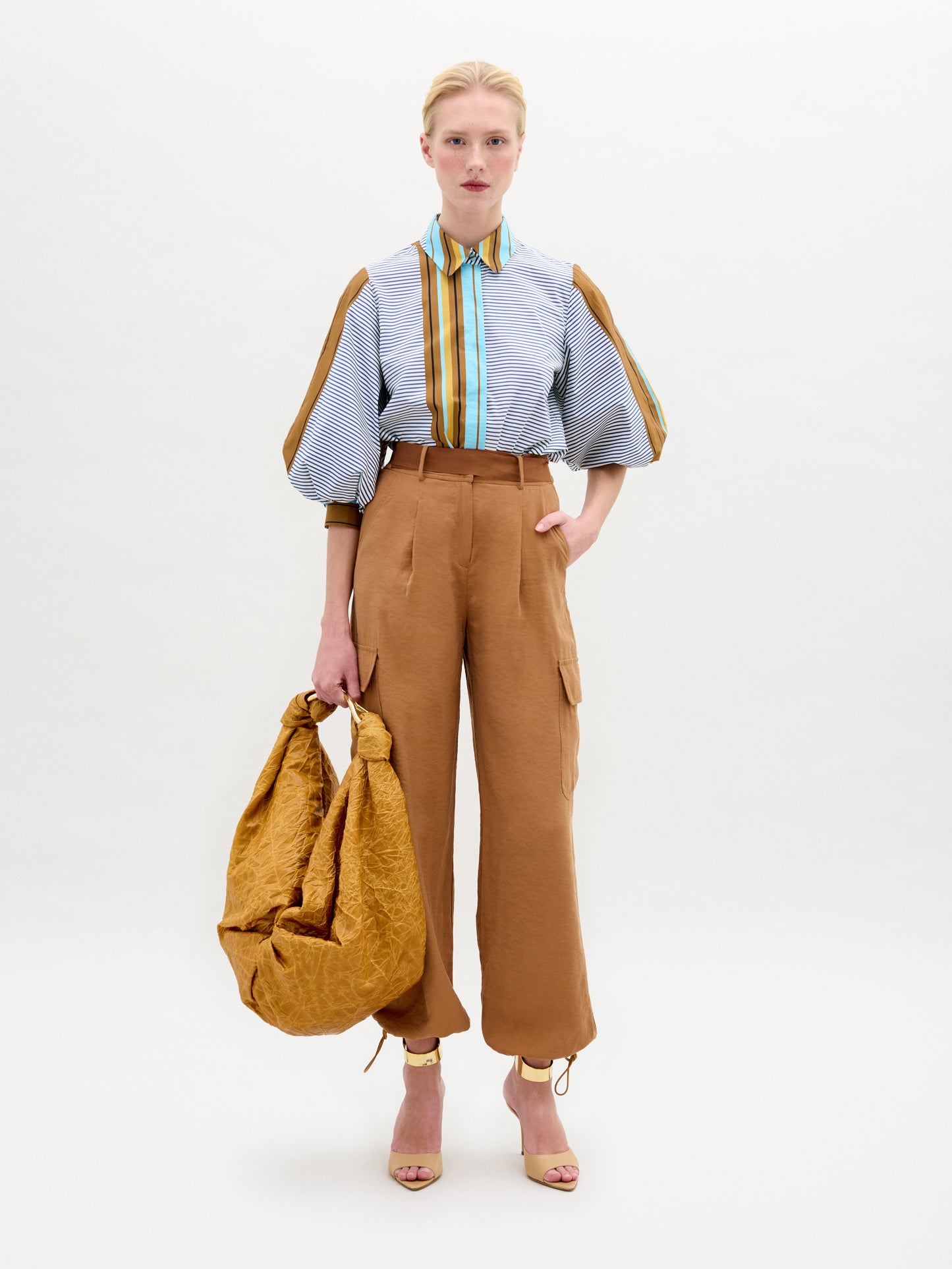 A person stands against a plain background wearing a blue striped shirt with puffy sleeves, SVT DESIGNS' Darnell Pant in relaxed-fit brown high-waisted style, and open-toed heels. They are holding a large, crumpled brown bag.