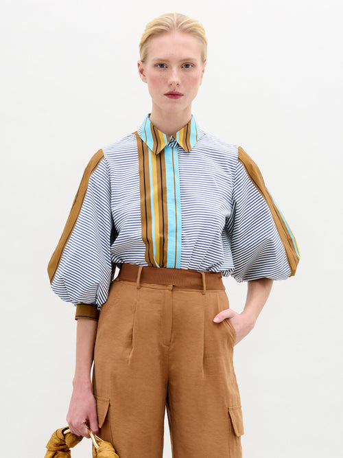 A person with blond hair is wearing the Belkis Blouse by SVT DESIGNS, featuring stripes and voluminous sleeves, paired with high-waisted oversized brown pants. They hold a gold-colored accessory against a plain white background.