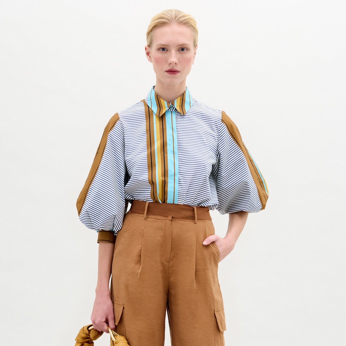 A person with light skin and blonde hair tied back wears the SVT DESIGNS Belkis Blouse, a loose cotton top with puffy sleeves, paired with oversized tan high-waisted pants. They hold a tan item in their left hand against a plain light background.