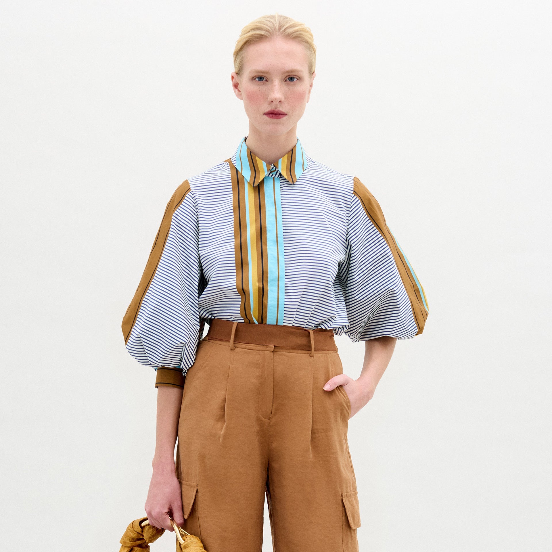 A person with light skin and blonde hair tied back wears the SVT DESIGNS Belkis Blouse, a loose cotton top with puffy sleeves, paired with oversized tan high-waisted pants. They hold a tan item in their left hand against a plain light background.