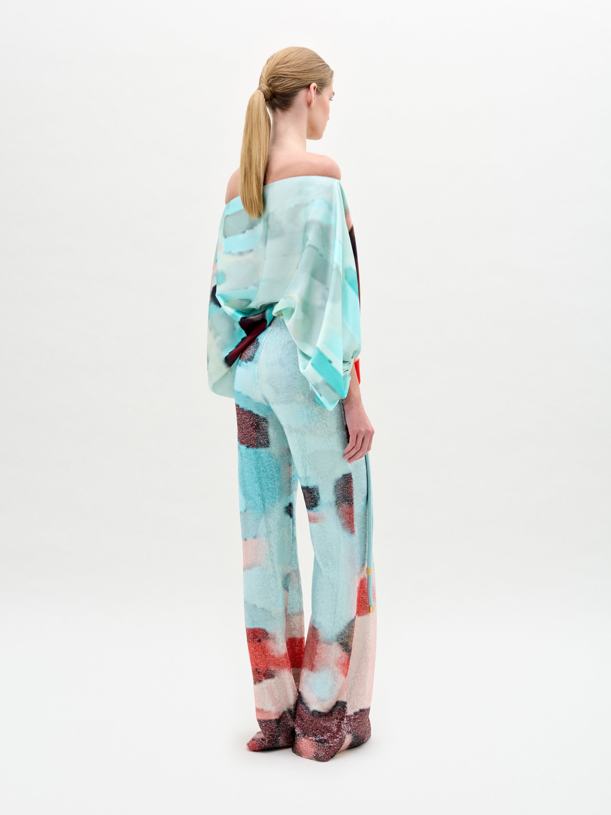 A person with blond hair in a low ponytail faces away, wearing SVT DESIGNS' Dalal Pant—high-waisted, wide-legged pants with a blue, white, and red pastel abstract pattern—paired with a loose off-the-shoulder top.