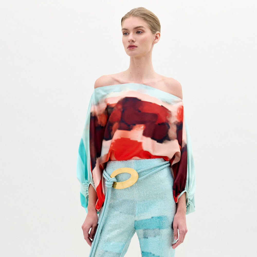 A woman stands against a plain background wearing the Bellagio Blouse by SVT DESIGNS—an off-the-shoulder, long-sleeve silk top with a red and aqua abstract pattern—paired with high-waisted light blue pants and a large beige belt buckle.