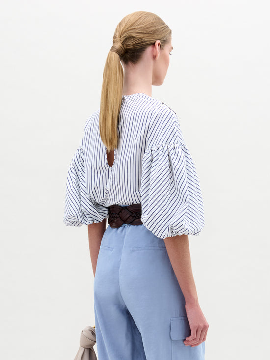 A person with long, tied-back hair is wearing the SVT DESIGNS Belle Blouse, a white and blue striped top made of luxurious Italian fabric with puffed sleeves, paired with light blue pants. They are holding a taupe-colored bag by their side while facing away from the camera.