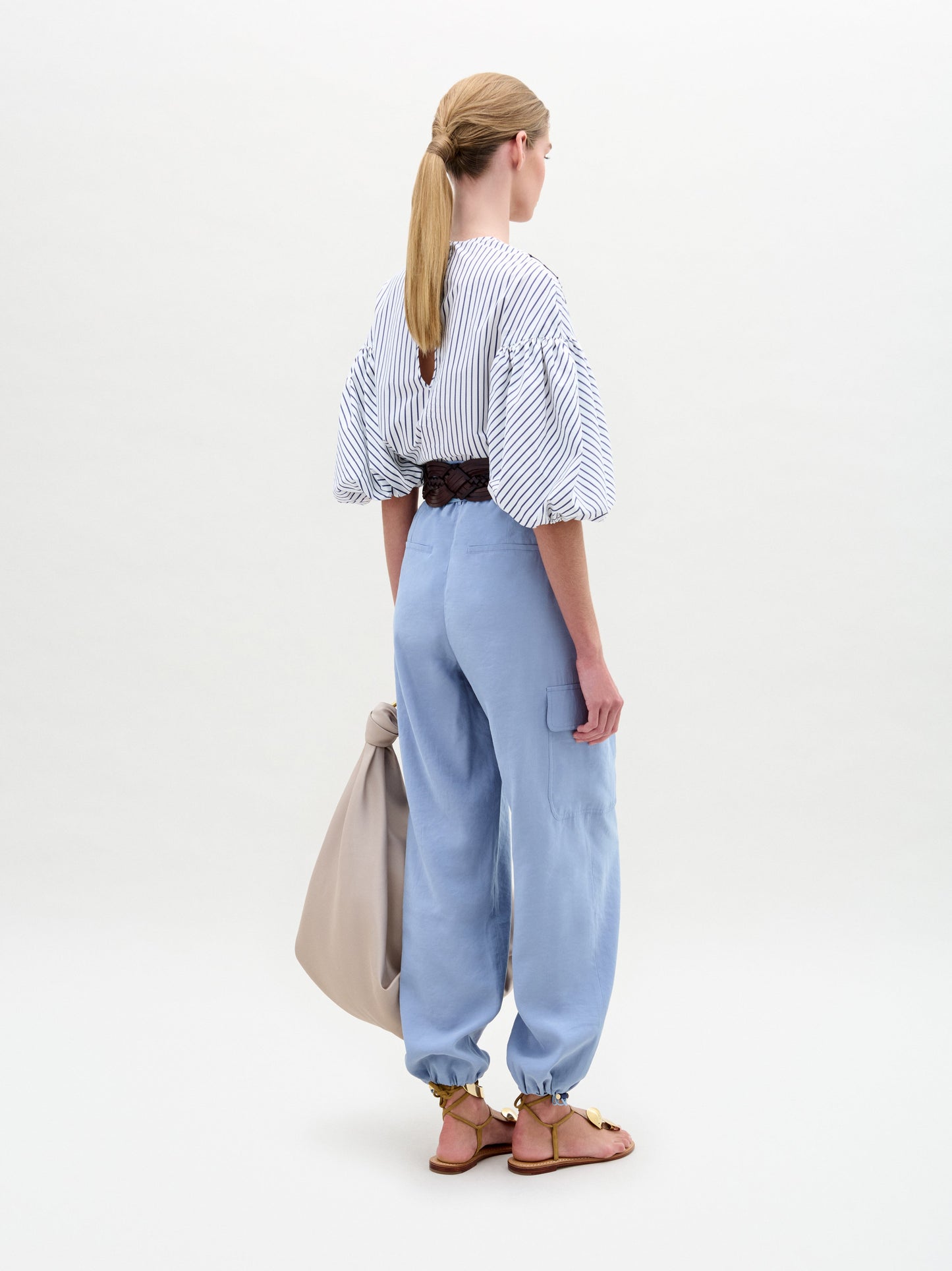 A person with blonde hair in a low ponytail sees wearing SVT DESIGNS' Darnell Pant, featuring high-waisted light blue with elastic cuffs and a striped shirt with puffy sleeves. Complete the look with brown sandals and a beige bag against a plain white backdrop.