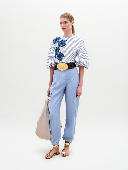 A woman stands against a plain white background wearing a striped blouse with blue floral embroidery, SVT DESIGNS' Darnell Pant in light blue Italian fabric, a wide belt, and strappy sandals. She holds a large beige bag at her side.