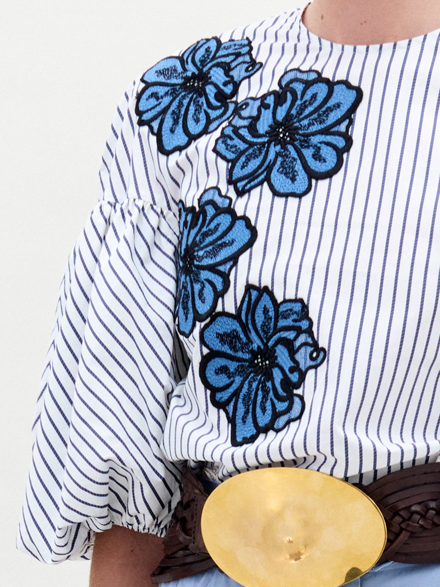 A person is wearing the SVT DESIGNS Belle Blouse, a 100% cotton white and blue striped blouse featuring blue floral embroidery on the shoulder and upper chest. It has puffed sleeves, accented by a wide dark belt with a large round gold buckle.