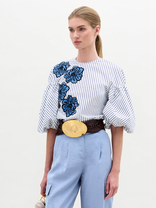 A person with long hair tied back is wearing SVT DESIGNS' Belle Blouse, a striped 100% cotton top with blue floral accents, light blue pants, and a wide belt with a large circular buckle. They are holding a small object in their left hand.