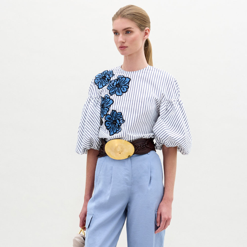 A woman stands against a plain background wearing the Belle Blouse by SVT DESIGNS—a white striped blouse with voluminous sleeves crafted from fine Italian fabric—paired with light blue trousers, a wide brown belt, and holding a small handbag.