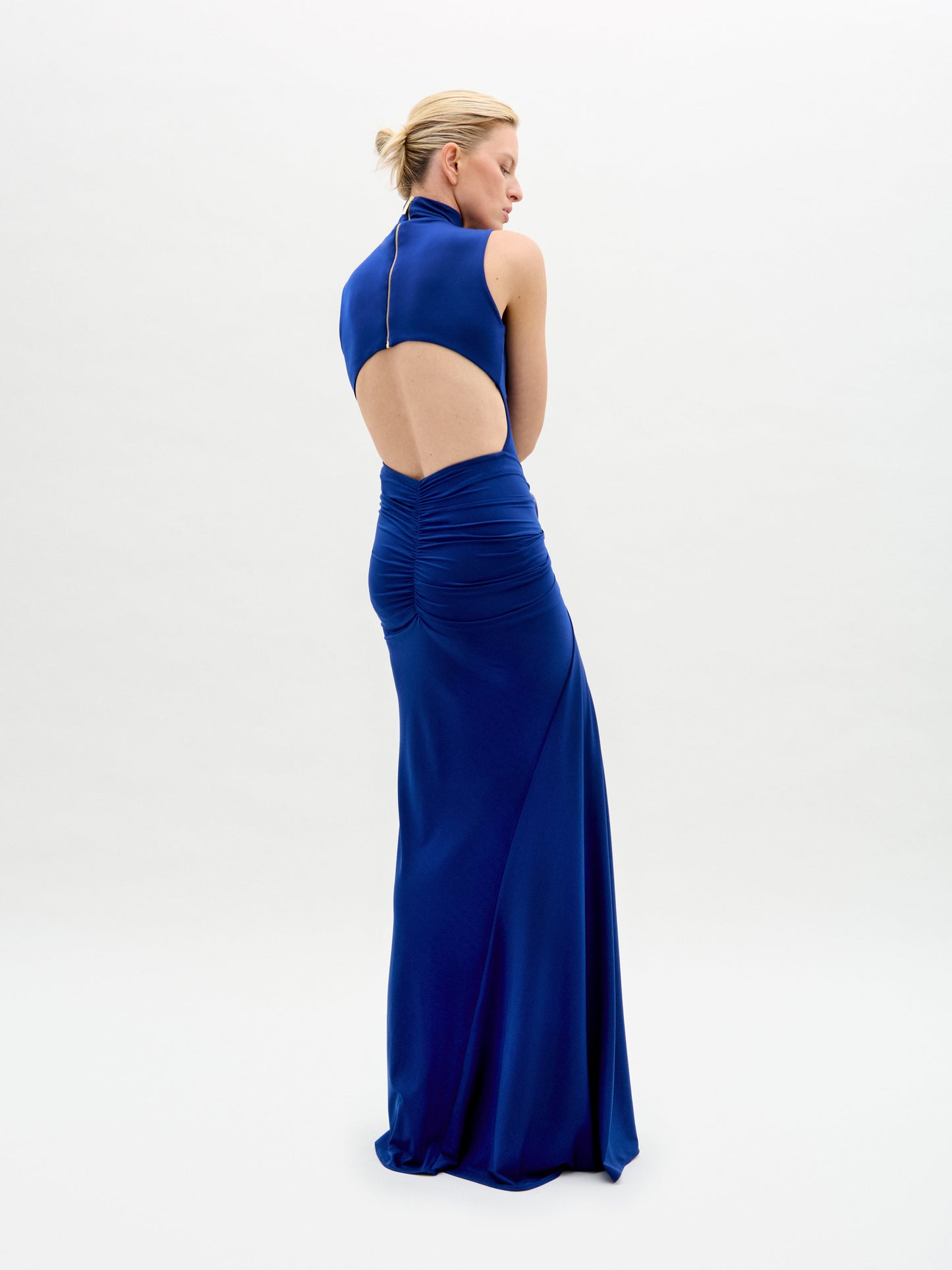 A woman with blond hair in an updo is posing against a plain white background wearing the Bogna Dress by SVT DESIGNS, a long sleeveless blue gown made from fine Italian fabric, featuring an open back and gathered detailing.