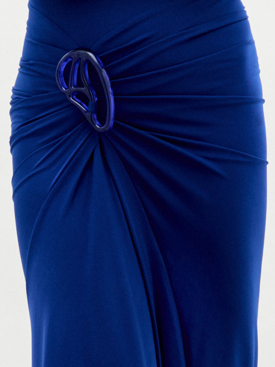 The Bogna Dress by SVT DESIGNS features a royal blue asymmetric skirt, highlighted by a gathered knot at the hip with a shiny abstract blue brooch. The Italian fabric drapes elegantly, creating smooth, flowing lines.