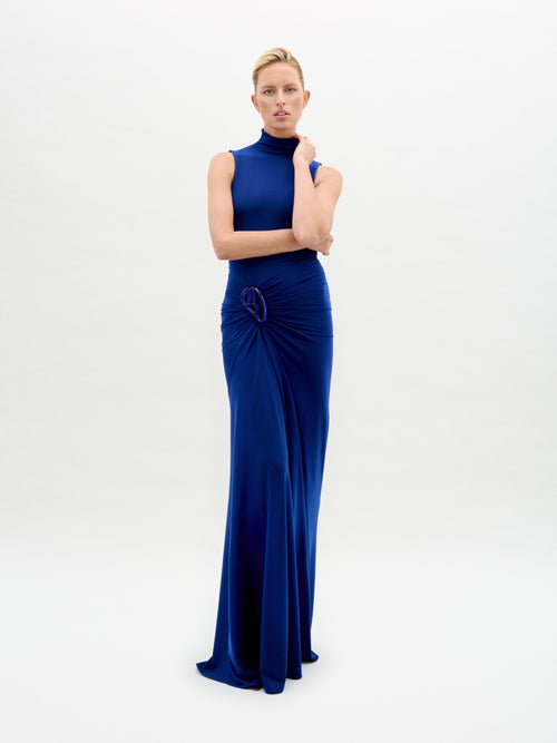 A person confidently poses against a plain background wearing the elegant, sleeveless high-necked Bogna Dress by SVT DESIGNS. Crafted from fine Italian fabric, it features a gathered waist with knot detail and an asymmetric skirt, while the person crosses their arms and faces the camera.