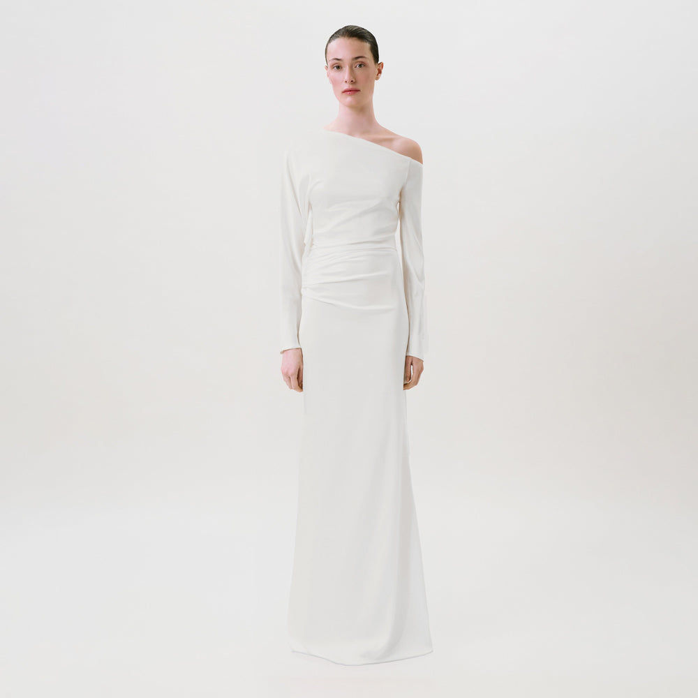 A woman stands against a plain white background wearing the Bahir Dress, a long, white, one-shoulder gown crafted from luxurious silk. This elegant ready-to-wear piece features long sleeves and a draped waist detail, expertly made in the USA for effortless sophistication.
