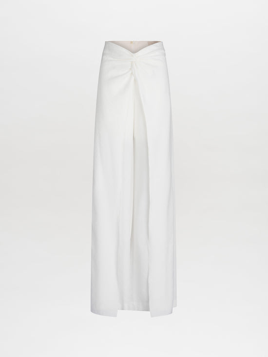 The Canturipe Pant by SVT DESIGNS is showcased against a plain backdrop, featuring Italian fabric in white. This high-waisted, wrap maxi skirt style includes a front pleat and drapes elegantly to the floor, embodying a clean and minimalist aesthetic.