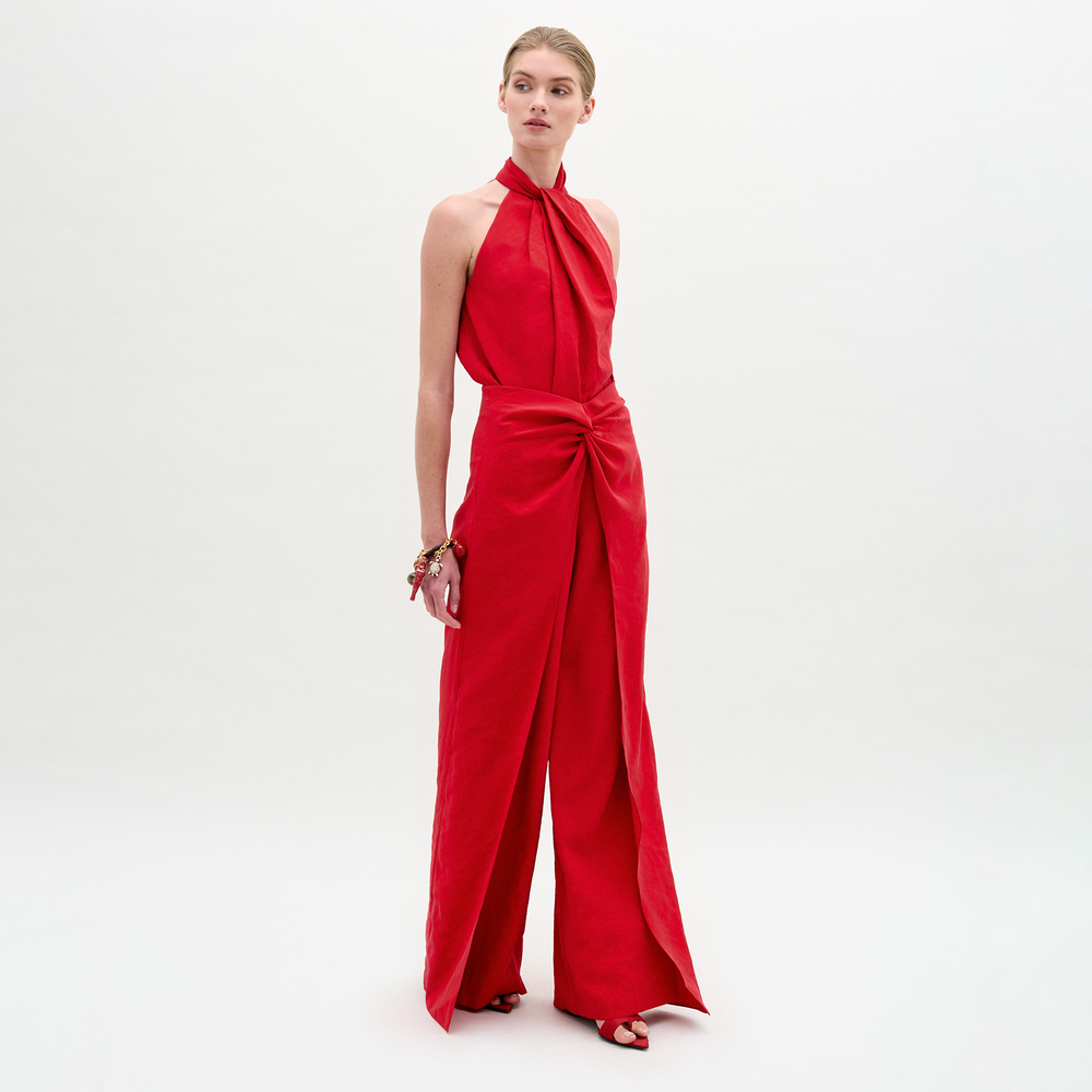A person stands against a white background wearing the Canturipe Pant by SVT DESIGNS, a high-waisted, wide-leg red Italian fabric pant with a twist detail at the waist, styled with pulled-back hair, bracelets, and red heeled sandals.