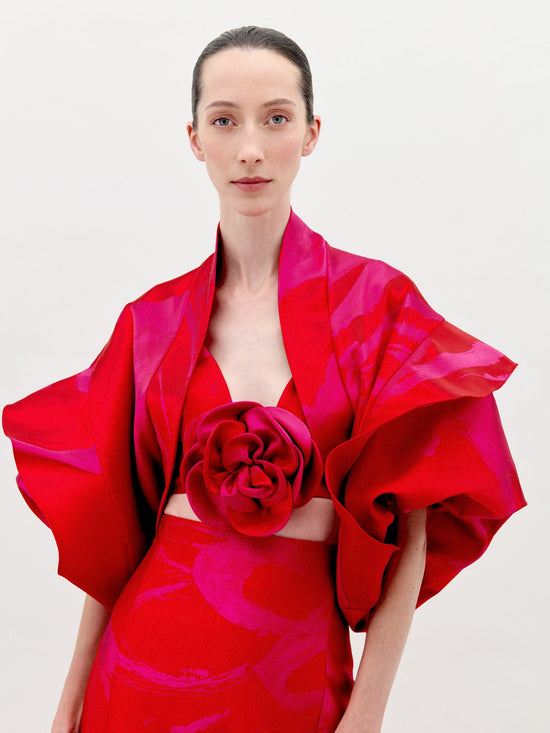 A person with fair skin and dark hair is wearing the Aleen Shrug Rouge Fuchsia Flower, a red, floral-themed outfit featuring large, petal-like sleeves and a central rosette. Set against a plain white background, this stunning piece is available for pre-order now and will ship by February 2025.