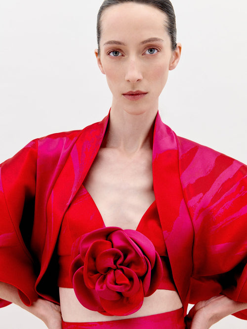 A person with fair skin and dark hair is wearing the Aleen Shrug Rouge Fuchsia Flower, a red, floral-themed outfit featuring large, petal-like sleeves and a central rosette. Set against a plain white background, this stunning piece is available for pre-order now and will ship by February 2025.