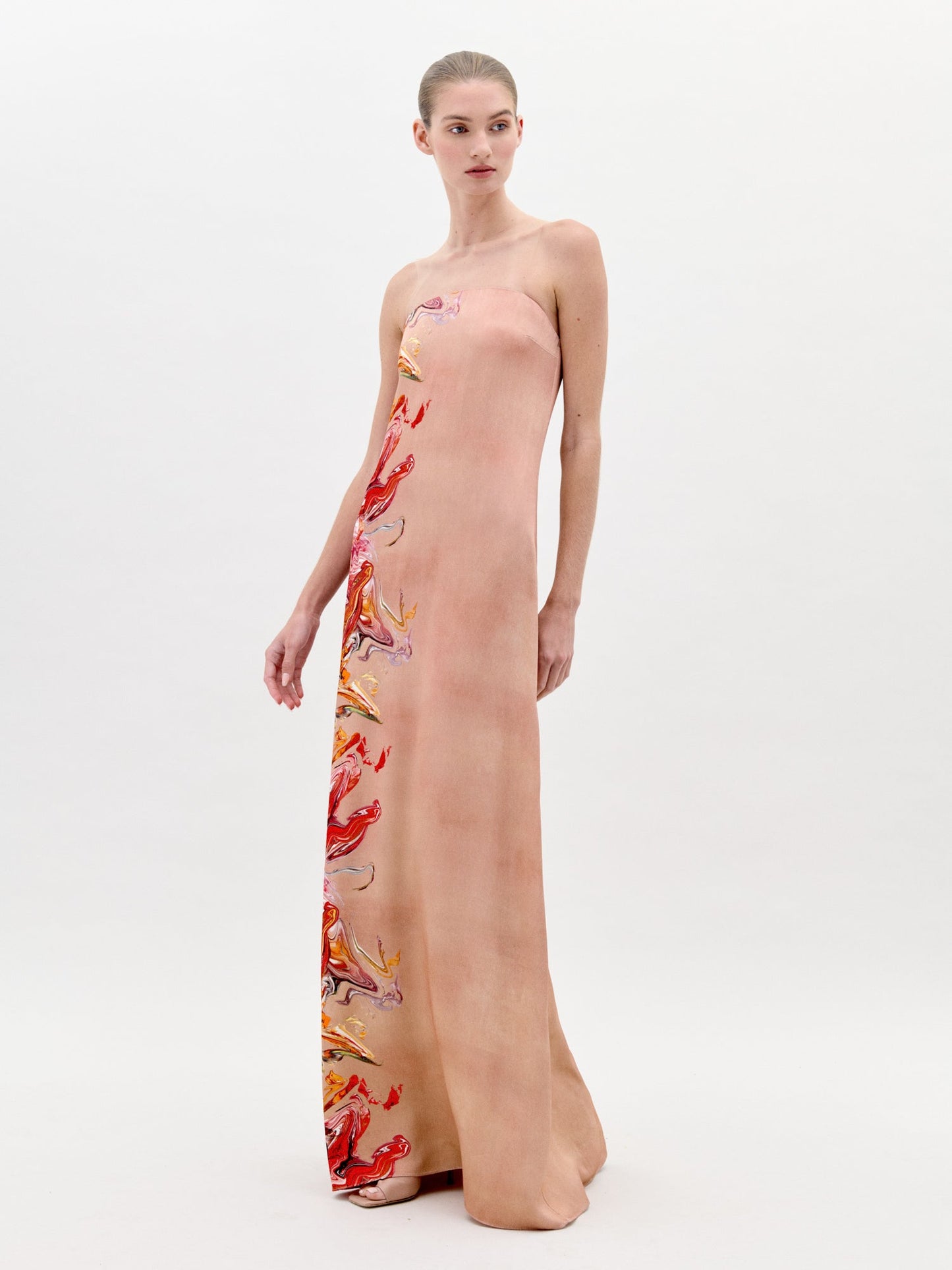 A woman stands in a long, strapless Adalgisa Dress Abstract Coral with red, orange, and yellow floral accents on the side against a plain white background. This stunning piece is available for pre-order now with an estimated ship-by date of February 1st, 2025.