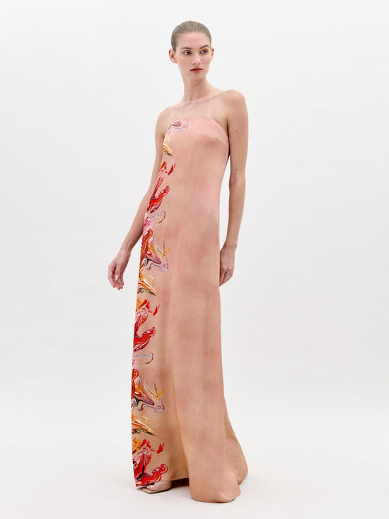 A woman stands in a long, strapless Adalgisa Dress Abstract Coral with red, orange, and yellow floral accents on the side against a plain white background. This stunning piece is available for pre-order now with an estimated ship-by date of February 1st, 2025.