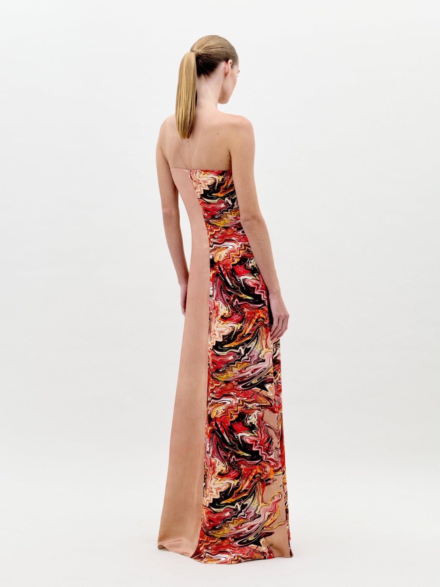 A woman stands in a long, strapless Adalgisa Dress Abstract Coral with red, orange, and yellow floral accents on the side against a plain white background. This stunning piece is available for pre-order now with an estimated ship-by date of February 1st, 2025.