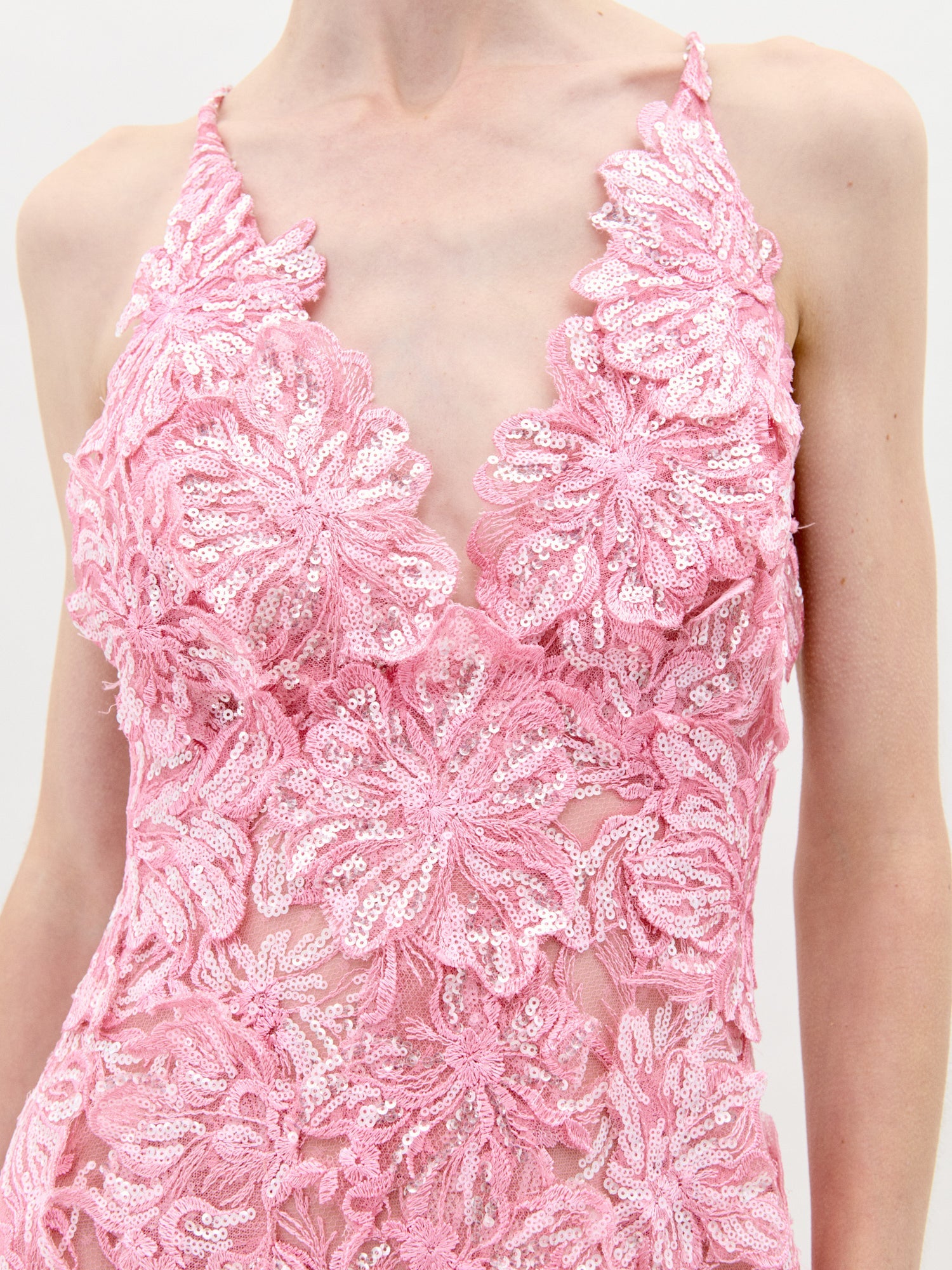 A person stands wearing the Daira Dress Pink, a long dress with thin straps and a beautiful floral pattern. The background is plain and light-colored. This elegant dress is now available for pre-order and will ship by February 1st, 2025.