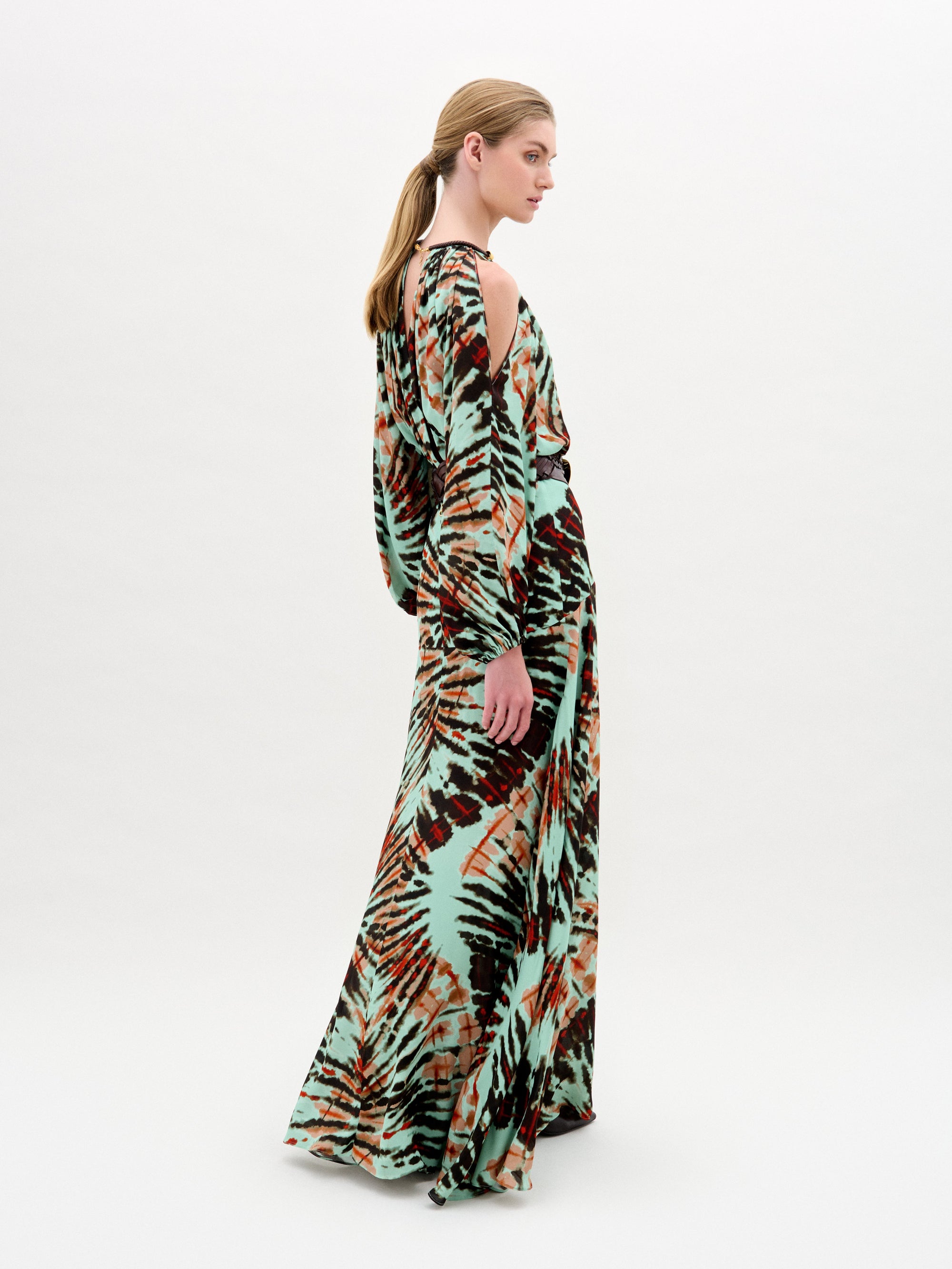 A woman poses in profile wearing the Carla Dress by SVT DESIGNS, a luxurious Italian-fabric maxi with a green, black, and brown multicolored tie-dye pattern; her hair is styled in a low ponytail against a plain background.