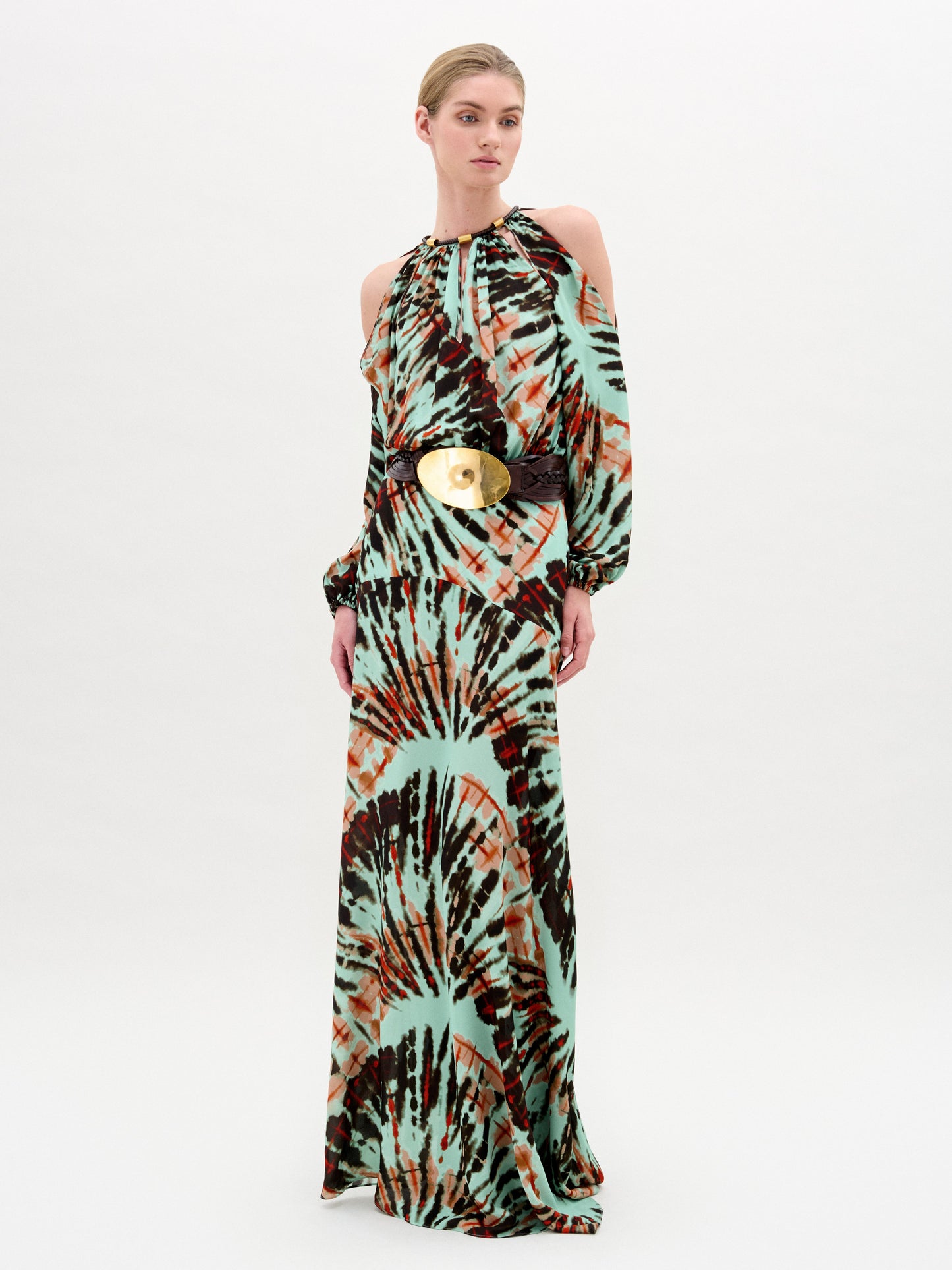 A woman models the Carla Dress by SVT DESIGNS, a luxe Italian maxi with green, brown, and orange tie-dye, high neckline, cold shoulder sleeves, and a wide belt with a large round gold buckle.
