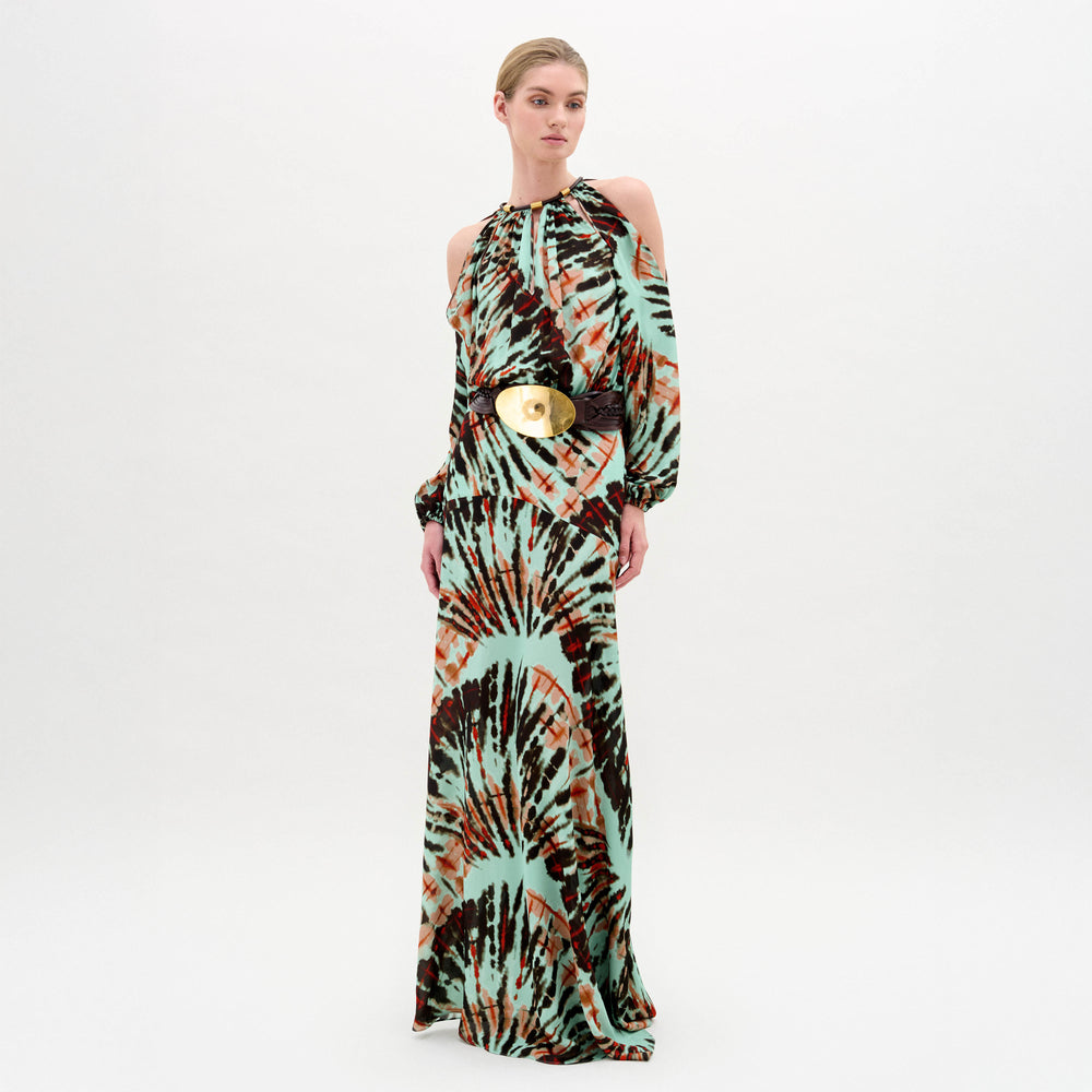 A woman poses against a white backdrop in the Carla Dress by SVT DESIGNS—a long, flowing maxi dress made from Italian fabric with a vibrant tie-dye pattern, shoulder cut-out long sleeves, and a bold black belt with a round gold buckle.