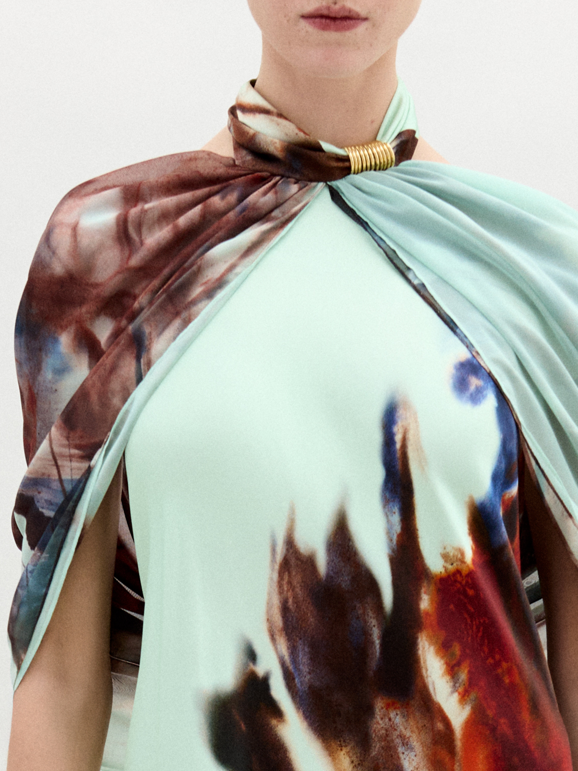 A woman stands against a white background wearing the Carlina Dress, a floor-length light blue maxi made from Italian fabric with a brown and red feather-like pattern and a matching draped shoulder cape.