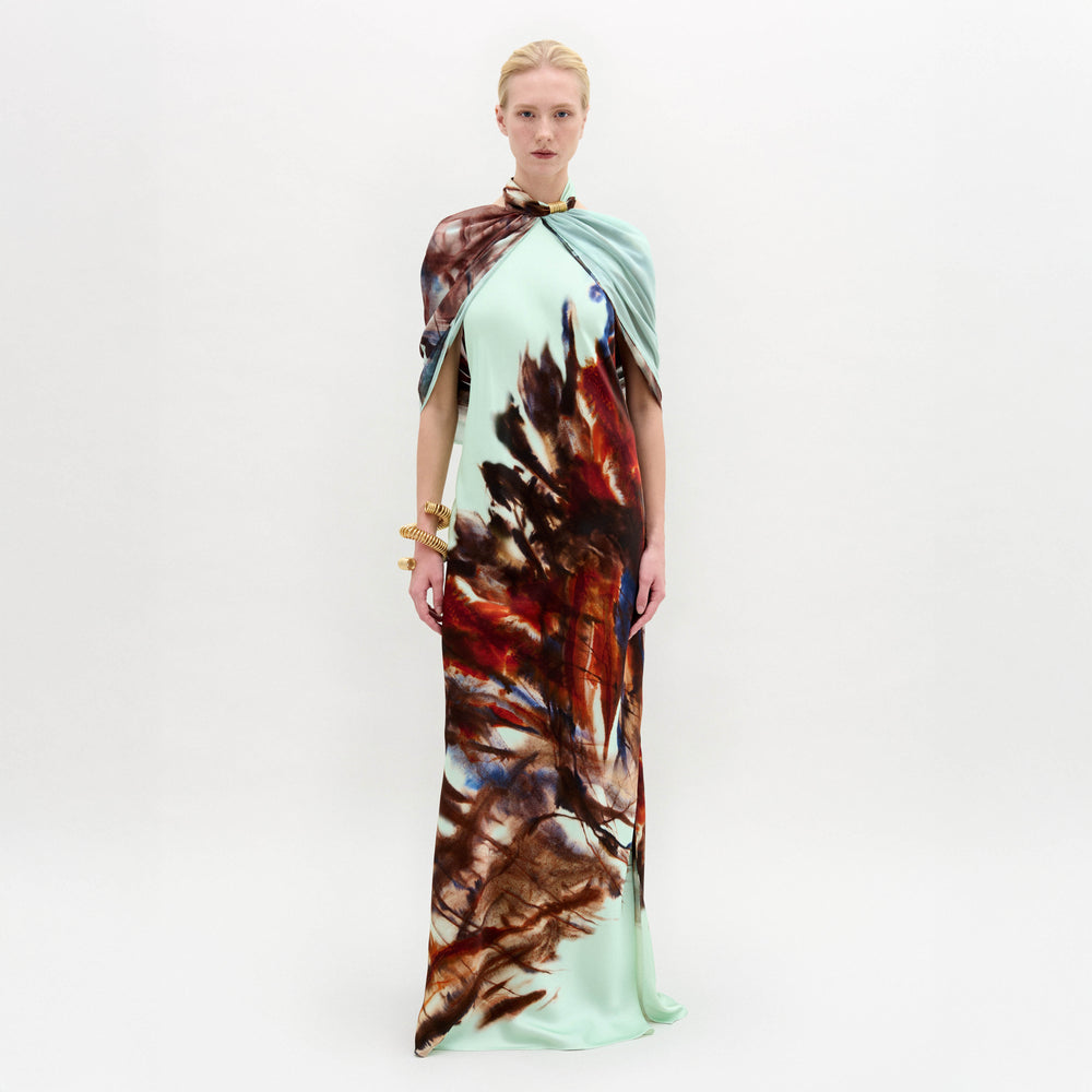 A person stands against a white background wearing the Carlina Dress by SVT DESIGNS—a floor-length Italian fabric mint green maxi with a brown and red feather-like pattern, matching draped cape, and gold bracelets.