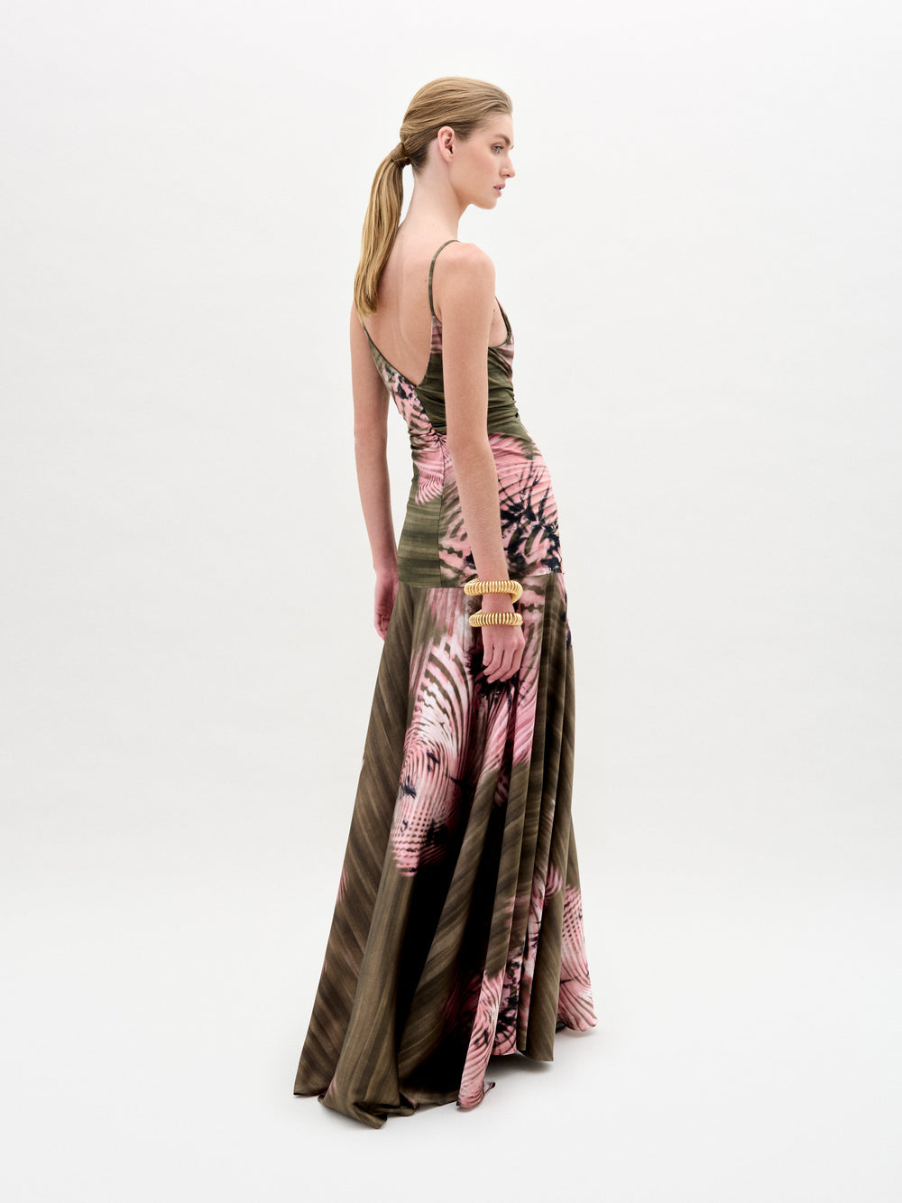A woman poses against a plain background wearing the Carmin Dress— a floor-length, sleeveless maxi crafted from Italian fabric with a pink and olive green tie-dye pattern and a deep V neckline.