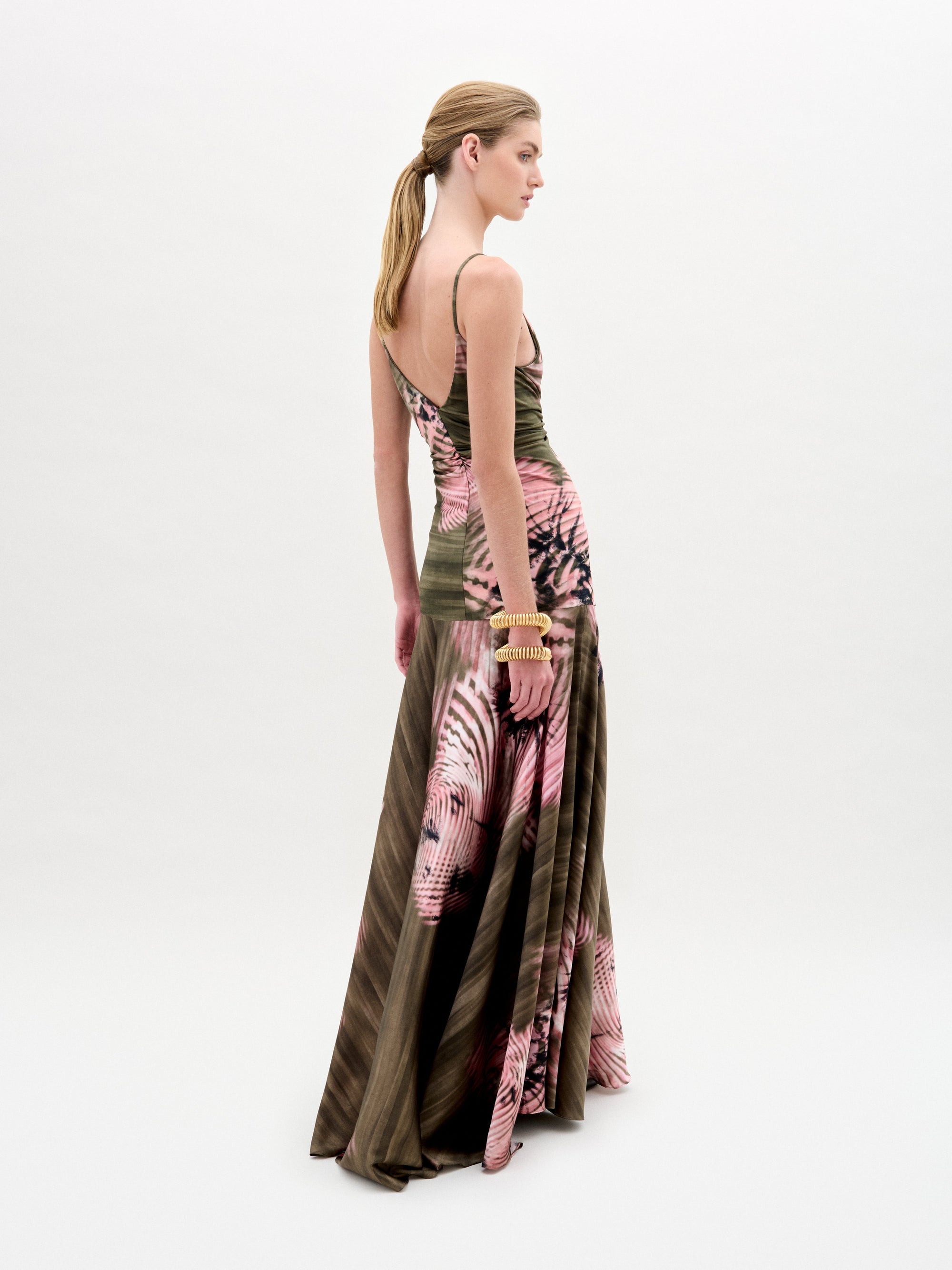 A woman poses against a plain background wearing the Carmin Dress— a floor-length, sleeveless maxi crafted from Italian fabric with a pink and olive green tie-dye pattern and a deep V neckline.