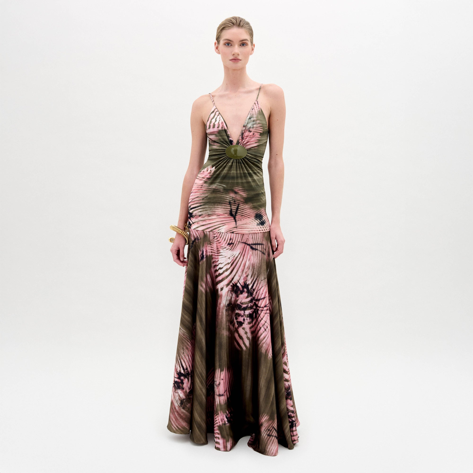 A woman poses against a plain background wearing the Carmin Dress— a floor-length, sleeveless maxi crafted from Italian fabric with a pink and olive green tie-dye pattern and a deep V neckline.