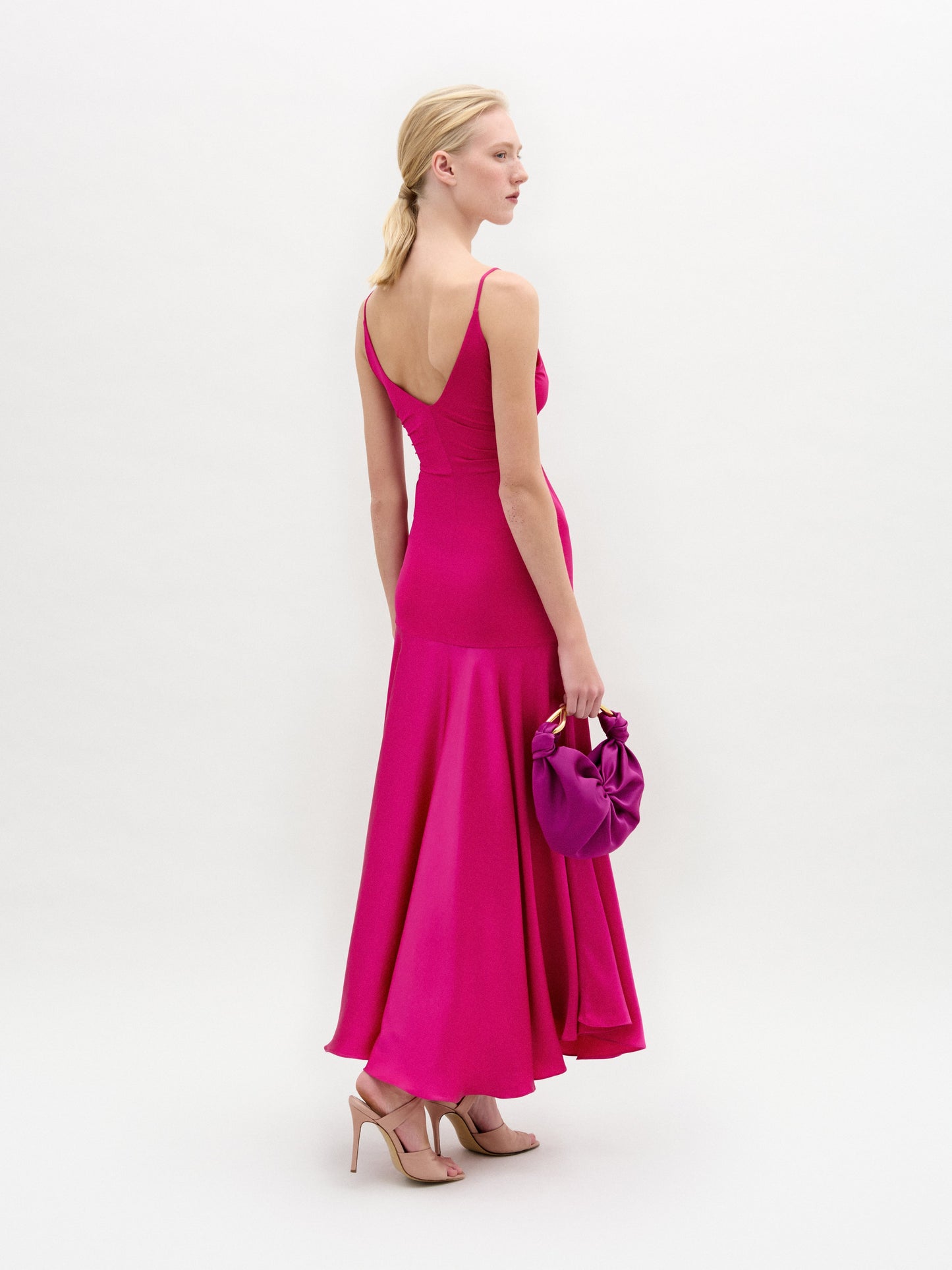 A woman poses against a white background in the Casiana Dress, a bright pink midi style made from Italian fabric with a deep V neckline. She pairs it with a matching small handbag and nude heels.