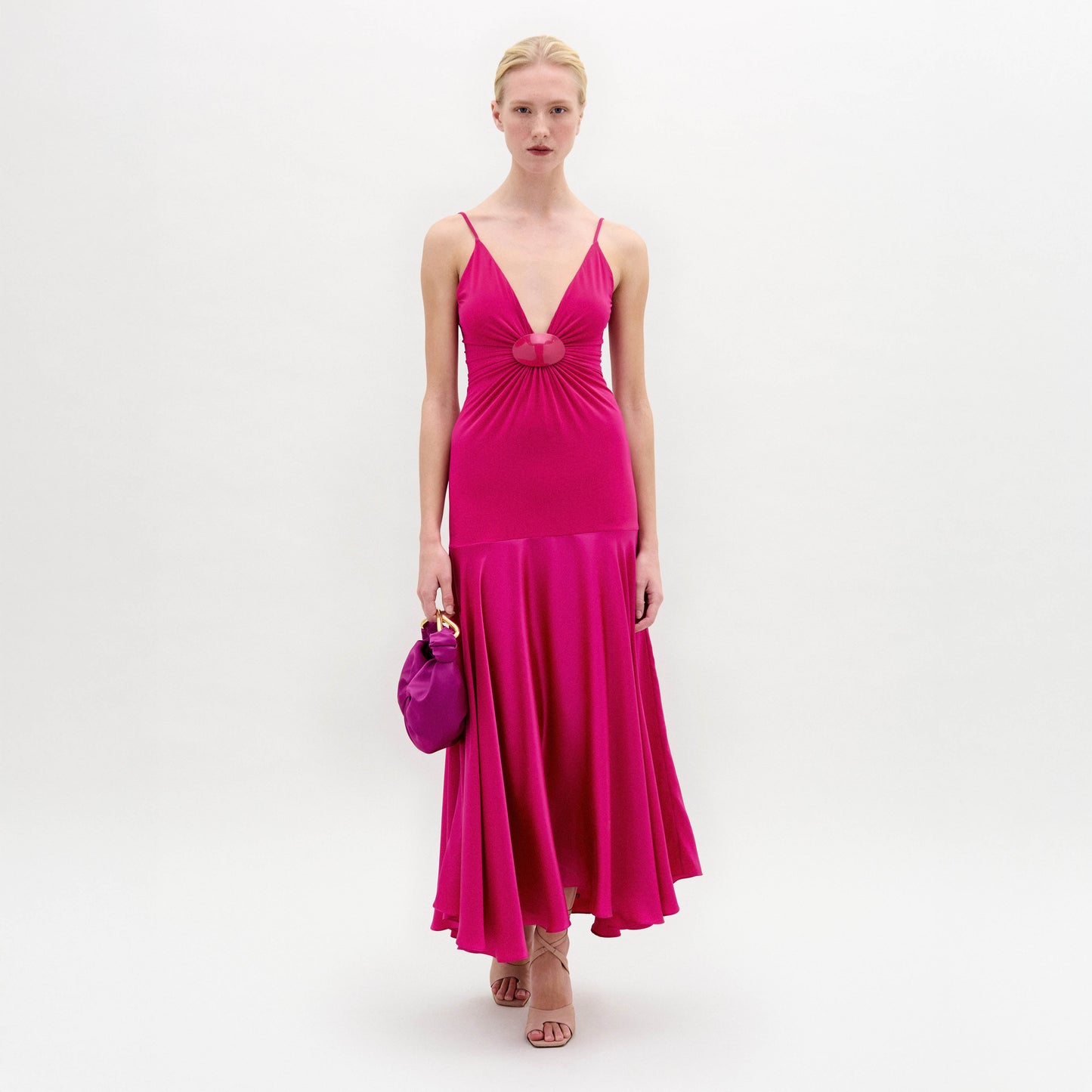 A woman poses against a white background in the Casiana Dress, a bright pink midi style made from Italian fabric with a deep V neckline. She pairs it with a matching small handbag and nude heels.