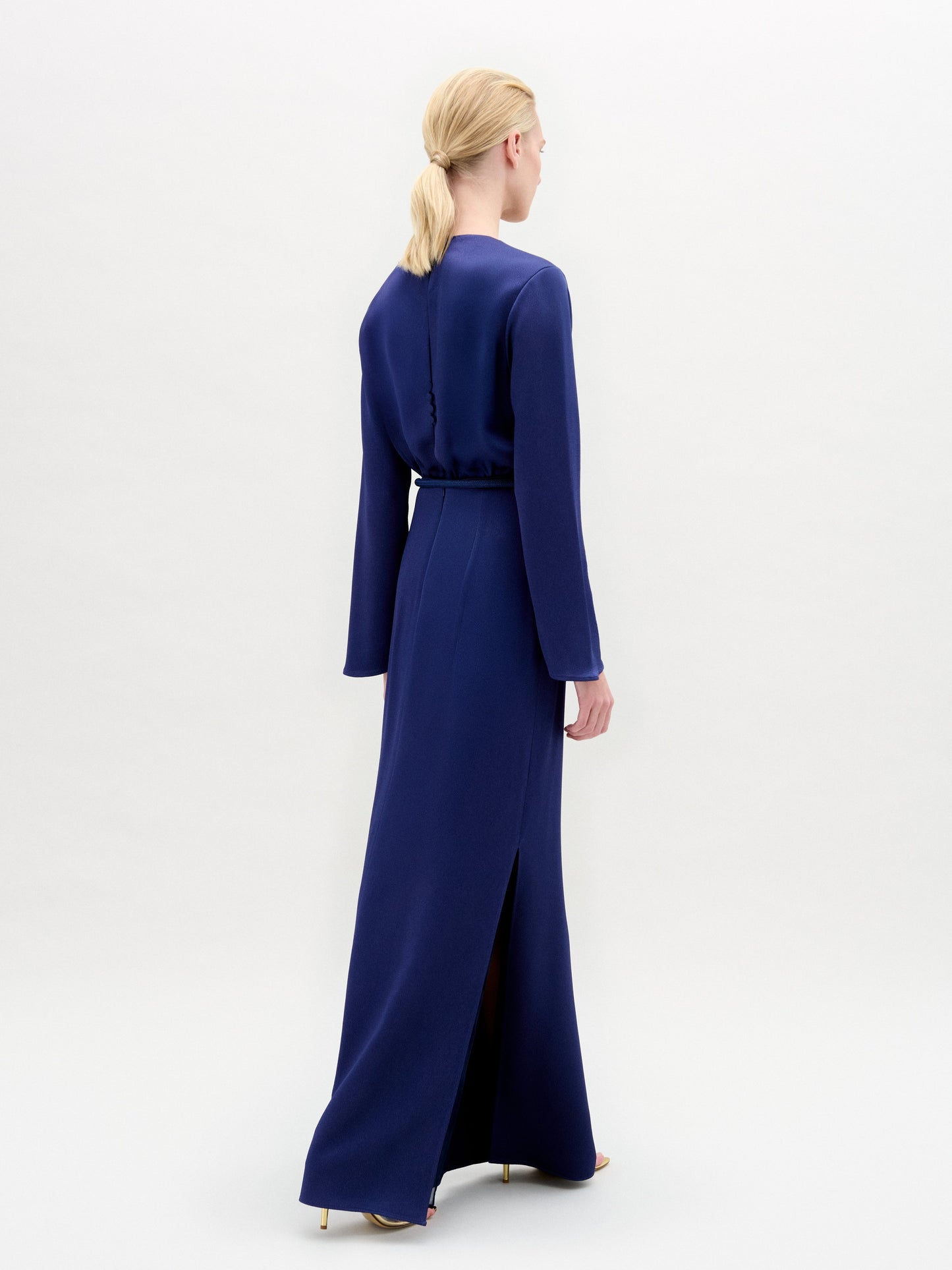 A person stands facing forward in the Cassy Dress—a long-sleeved, floor-length dark blue maxi with a keyhole neckline and black belt, crafted from luxurious Italian fabric—set against a plain white background.