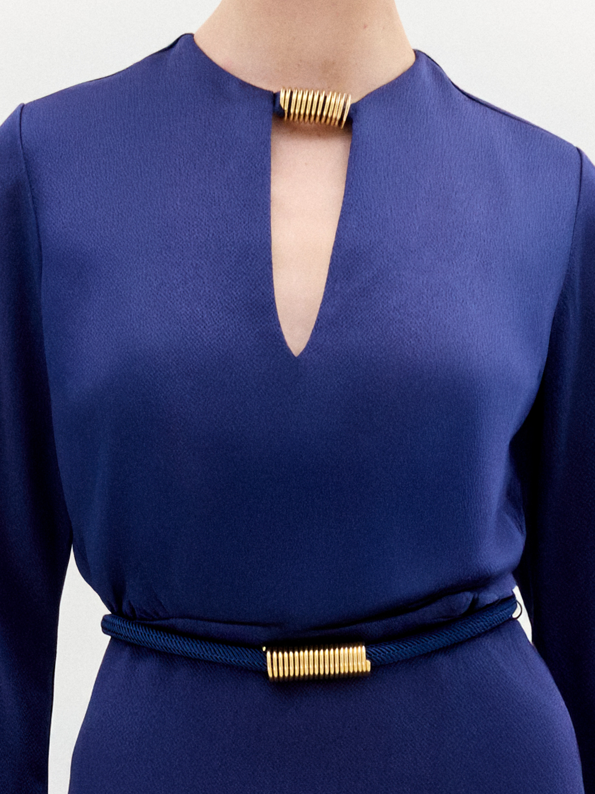 A person stands facing forward in the Cassy Dress—a long-sleeved, floor-length dark blue maxi with a keyhole neckline and black belt, crafted from luxurious Italian fabric—set against a plain white background.