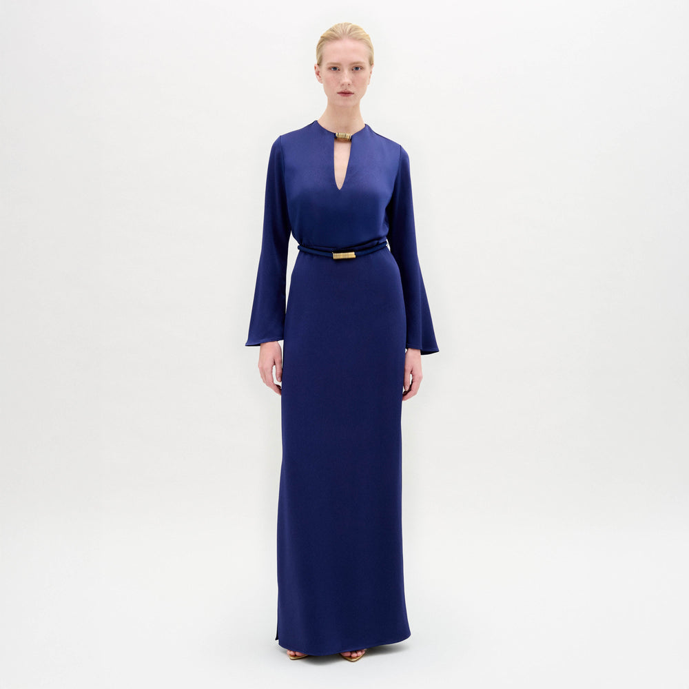 A person stands facing forward in the Cassy Dress—a long-sleeved, floor-length dark blue maxi with a keyhole neckline and black belt, crafted from luxurious Italian fabric—set against a plain white background.