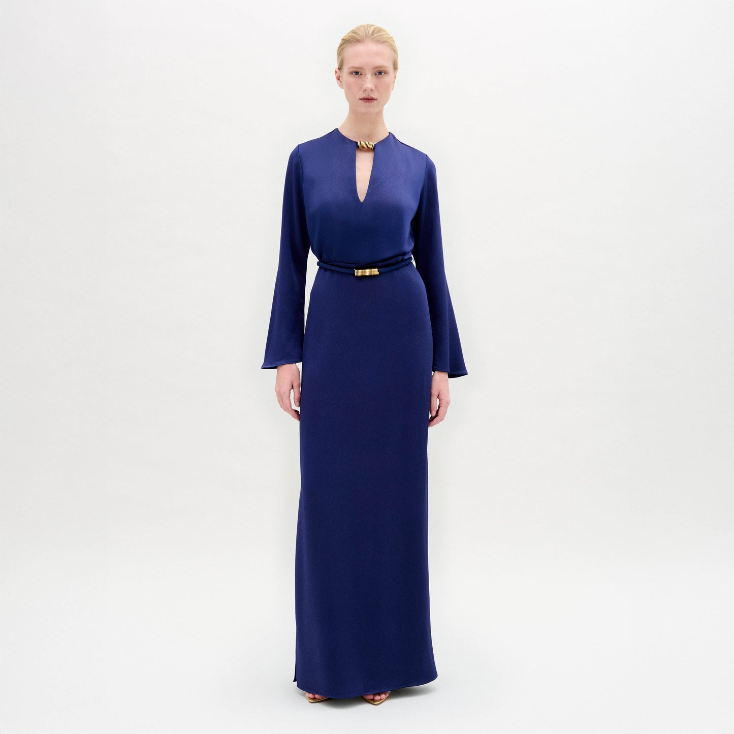 A person stands facing forward in the Cassy Dress—a long-sleeved, floor-length dark blue maxi with a keyhole neckline and black belt, crafted from luxurious Italian fabric—set against a plain white background.
