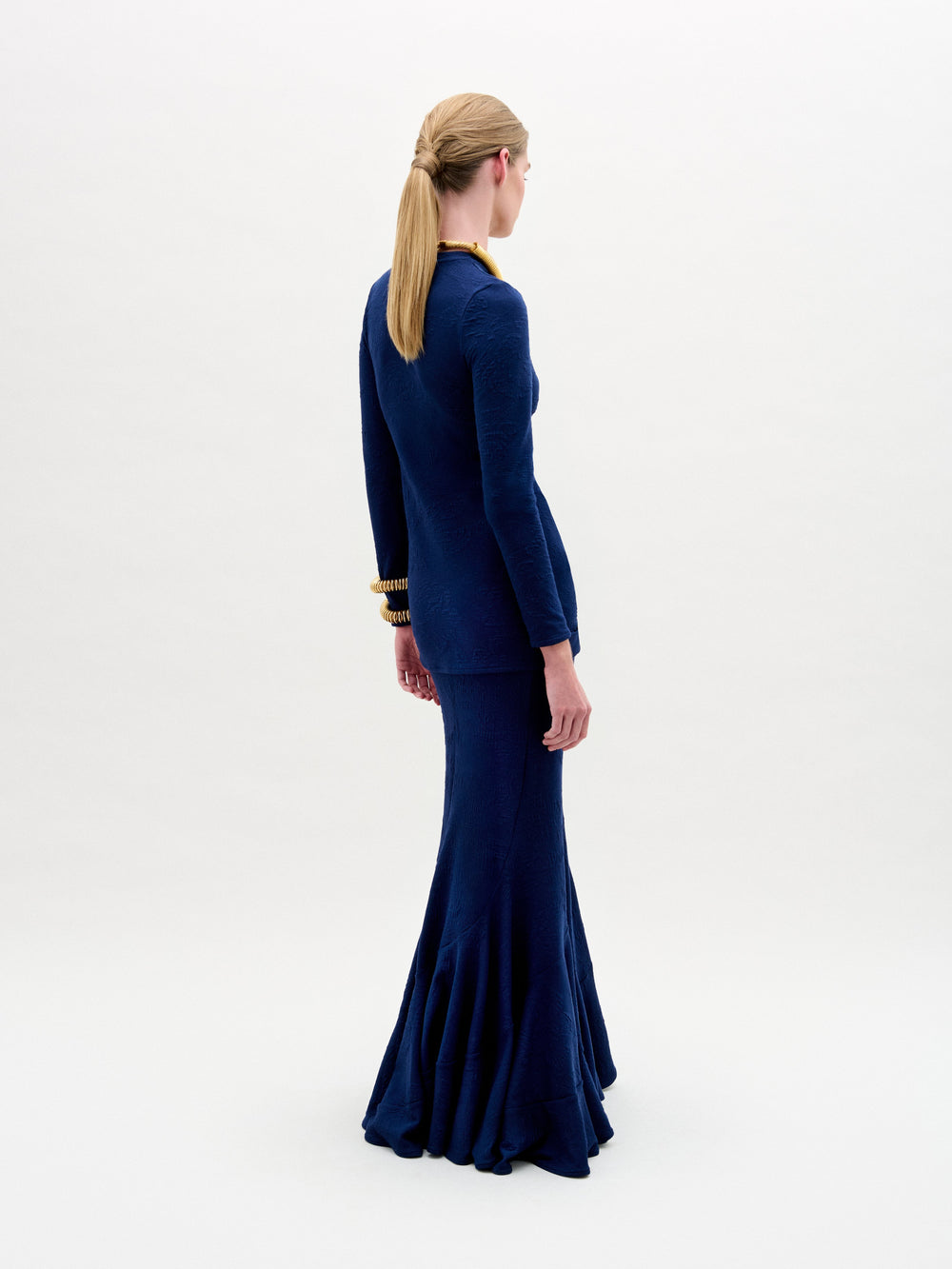 A person is against a plain background, wearing the Florina Skirt by SVT DESIGNS. The fitted dark blue dress has long sleeves and a high waist with a flared skirt. Their hair is styled in a low ponytail, complemented by gold bracelet and necklace accents.