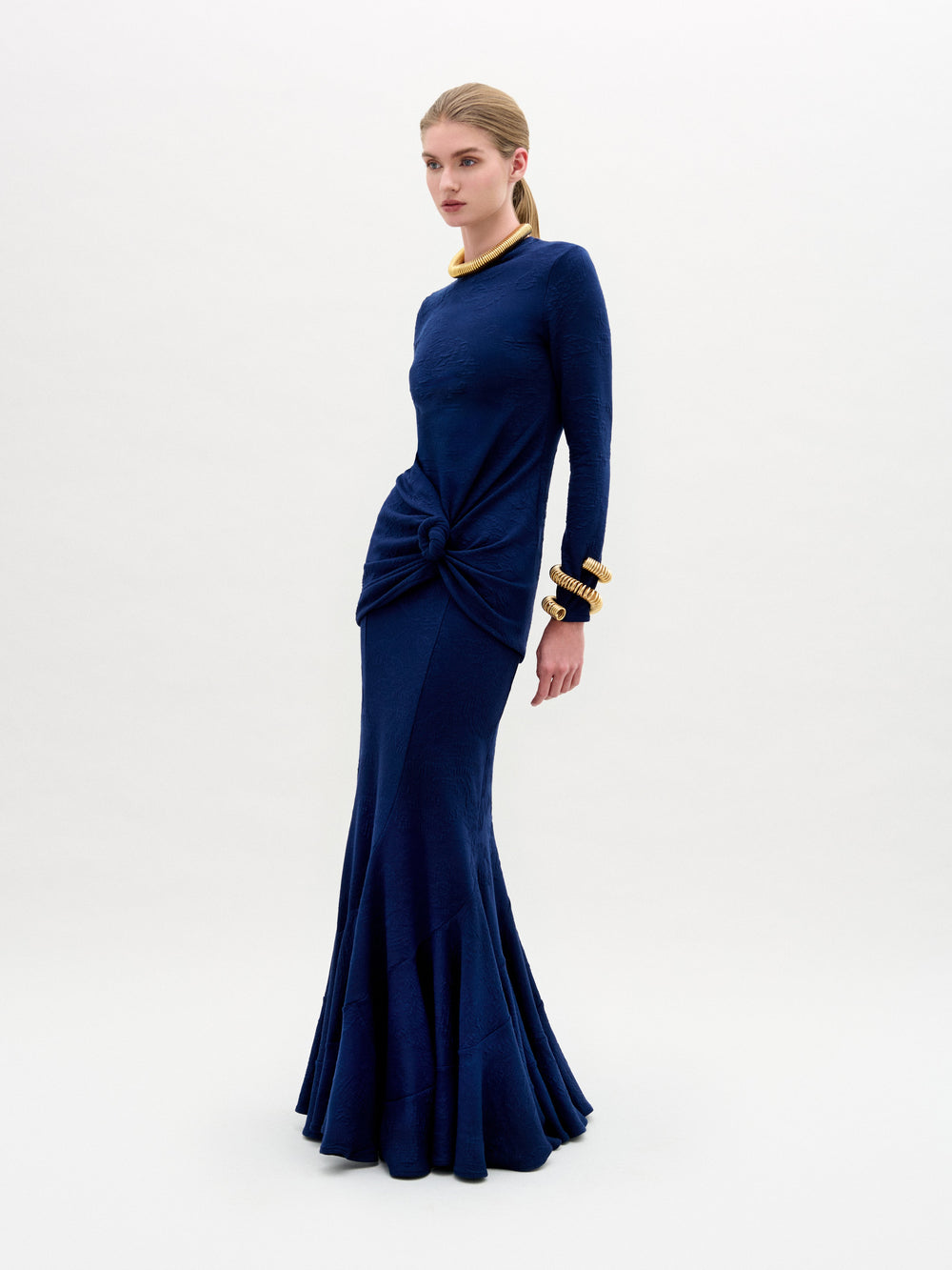 Against a plain white backdrop, a woman models the SVT DESIGNS Florina Skirt, rich navy blue and full-length with luxurious Italian fabric and knot detail at the waist. Her ponytail, gold bracelets, and necklace enhance the elegance of her outfit.