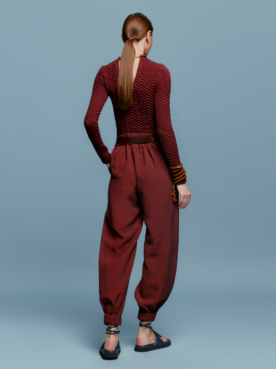 A person is standing against a blue background, wearing a textured maroon long-sleeved top, the Chilean Pant Burgundy with a belt, and open-toe sandals, evoking the casual elegance one might wear while boarding a ship for an August 19th departure.