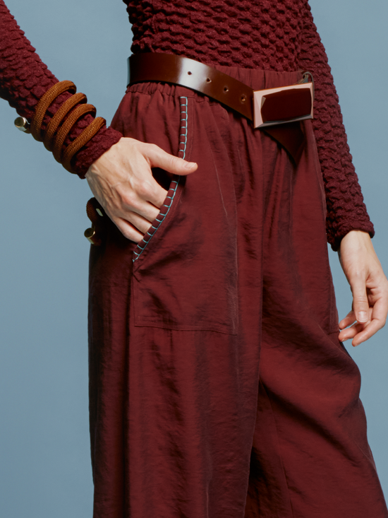 A person is standing against a blue background, wearing a textured maroon long-sleeved top, the Chilean Pant Burgundy with a belt, and open-toe sandals, evoking the casual elegance one might wear while boarding a ship for an August 19th departure.