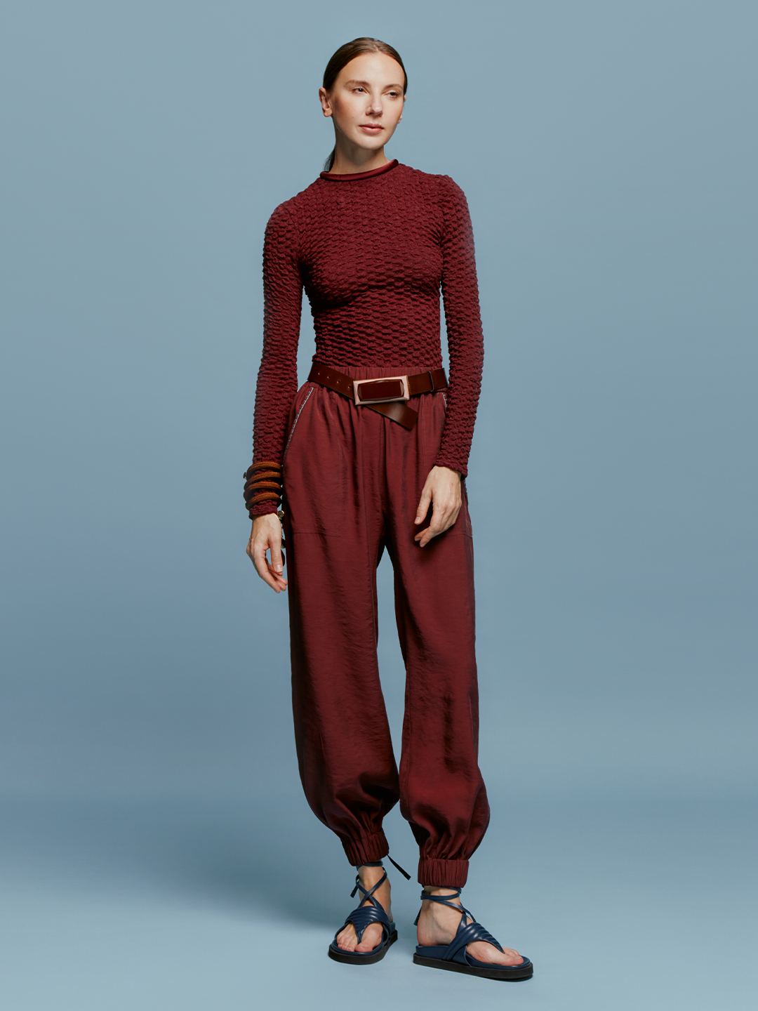 A person is standing against a blue background, wearing a textured maroon long-sleeved top, the Chilean Pant Burgundy with a belt, and open-toe sandals, evoking the casual elegance one might wear while boarding a ship for an August 19th departure.